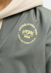 Šedá mikina s kulatým výstřihem, která má velký žlutý grafický potisk s nápisem "JACK&JONES 1990 URBN COLLECTORS CREW" na hrudi. Hladká textura.