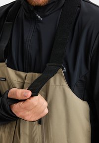 Bež vodoodporni kombinezon z nastavljivimi črnimi trakovi, teksturiran material in žep na zadrgo. Nosi se čez črno dolgodlako majico.