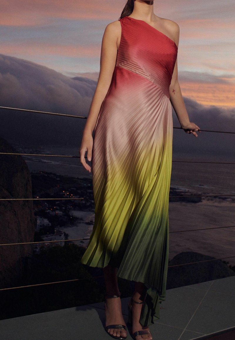 Femme portant une robe plissée sans manches à une épaule avec un dégradé rouge, jaune et vert, debout près d'une rambarde au coucher du soleil avec vue sur un paysage côtier.