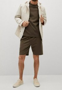 Giacca beige leggera con cappuccio, abbinata a una maglietta marrone scuro a maniche corte e pantaloni corti coordinati. Presenta scarpe da ginnastica color sabbia.