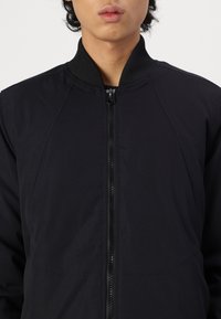 Black bomber jacket with a zipper front, ribbed collar, and smooth texture. Features structured seams for a modern fit.