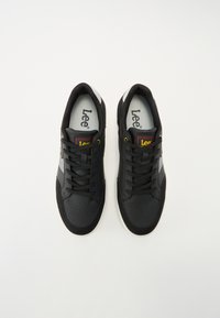 Baskets noires avec une tige en cuir lisse, des accents gris, un bout perforé et des lacets noirs. Présente un logo rouge et jaune sur la languette.