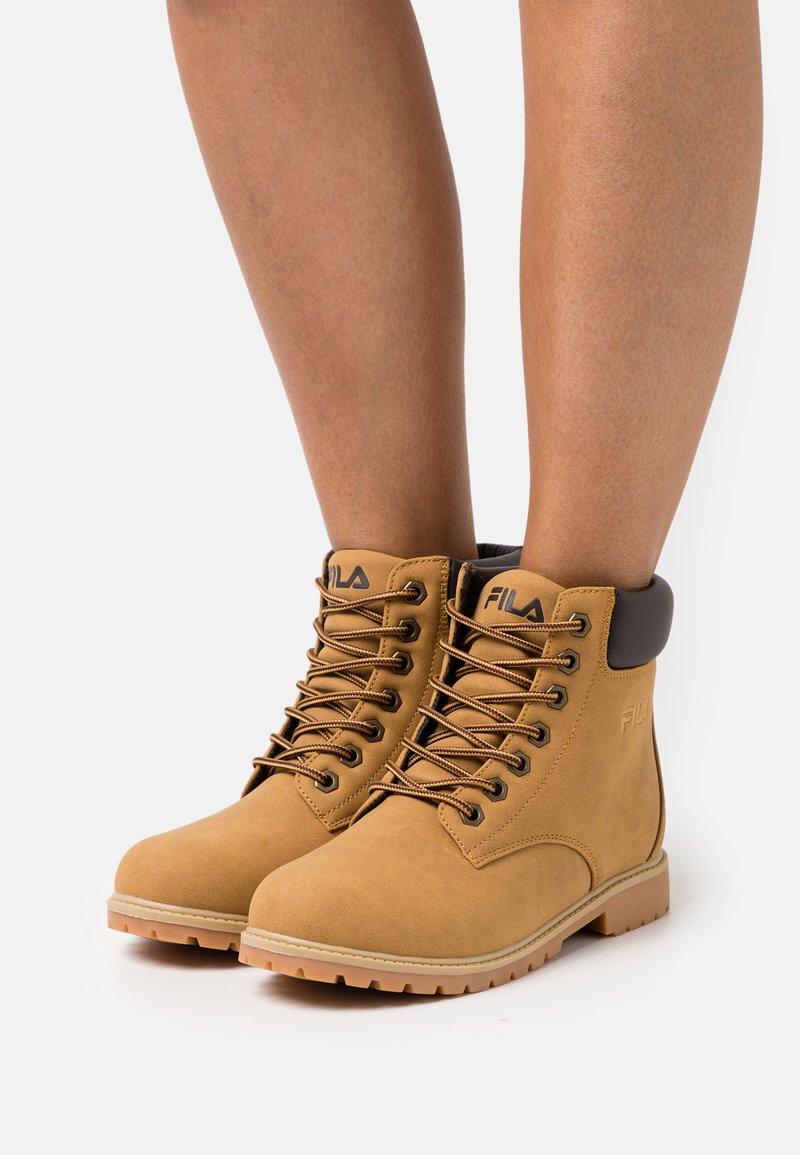 Bottines montantes marron clair FILA à lacets avec col matelassé noir portées sur jambes nues sur fond blanc.