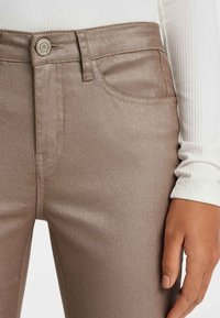 Jean en bronze métallique avec une texture lisse, design à cinq poches et fil assorti. Doté d'une fermeture à bouton rond et d'une silhouette ajustée.