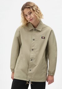 Lys beige jakke med krage, frontknapper, elastiske mansjetter og sidelommer; har et logo-merke på brystet.