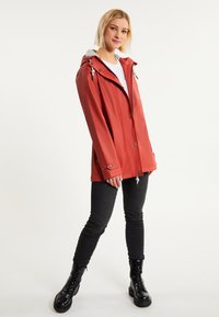 Chaqueta impermeable roja con capucha, con herrajes plateados, bolsillos frontales y un corte ligeramente ajustado. Lleva una blusa blanca y jeans oscuros.