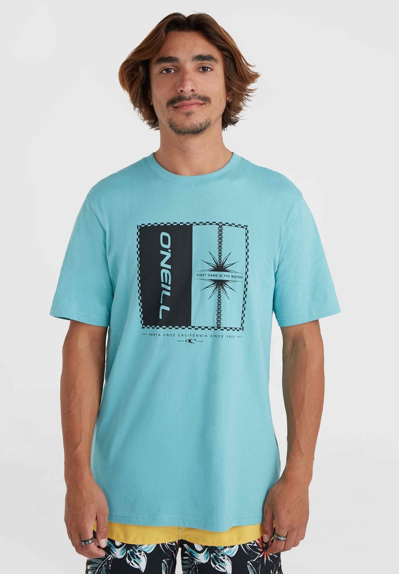 Camiseta de manga corta turquesa con un gráfico central. El diseño presenta acentos en negro, un logo y formas geométricas. Material de algodón.