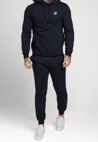 Uomo con felpa nera aderente e pantaloni sportivi abbinati con logo bianco, indossando scarpe bianche, in piedi contro uno sfondo semplice.