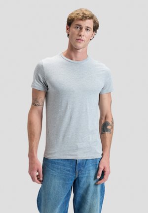 Junger Mann mit kurzen lockigen Haaren, trägt ein hellgraues T-Shirt und blaue Jeans, steht vor einem einfarbigen hellen Hintergrund.
