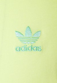 Un tessuto giallo chiaro presenta un logo Adidas ricamato in verde con un design simile a una foglia sopra il nome del marchio, su una superficie liscia.