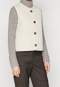 Krémová prošívaná vesta s přiléhavým střihem, vybavená pěti dřevěnými knoflíky, bočními kapsami a pod ní měkký, texturovaný pletený svetr.