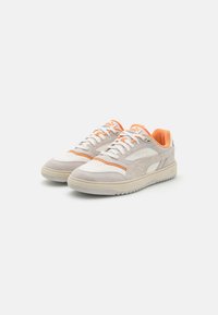 Une paire de baskets basses en suède beige, avec des panneaux en cuir blanc, des accents orange et des lacets, inclinée à droite sur un fond blanc.
