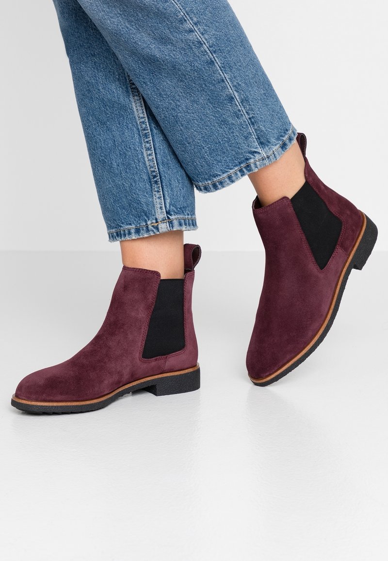 Bottines Chelsea en daim bordeaux montant à la cheville avec panneaux élastiques latéraux noirs, portées avec un jean bleu retroussé sur un sol blanc.