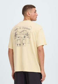 T-shirt en coton jaune clair avec un graphique au dos représentant des montagnes, des arbres et deux personnes assises sur des chaises. Manches courtes, coupe décontractée.