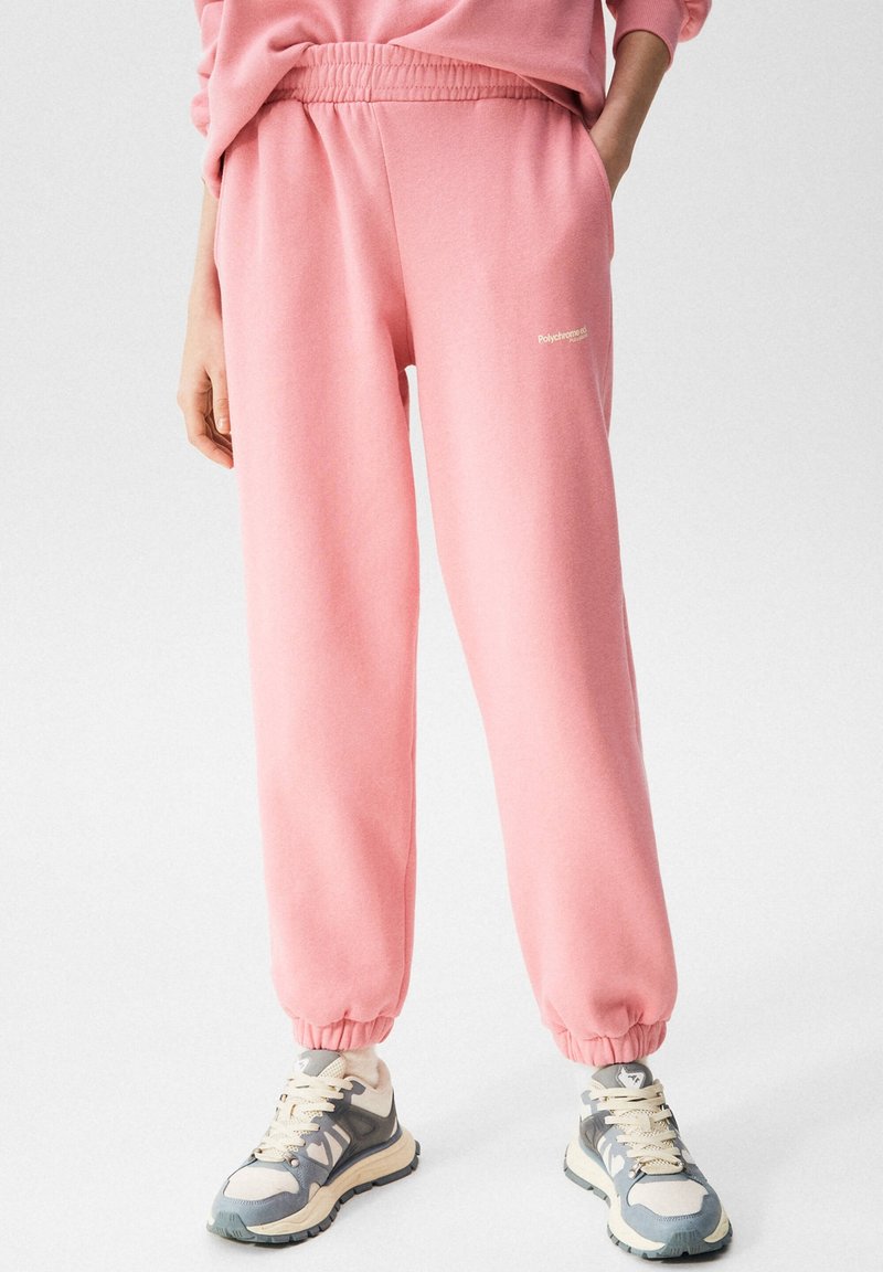 Bas du corps portant un pantalon de survêtement rose ample avec des poignets élastiques et des poches, assorti à des baskets épaisses grises et blanches.