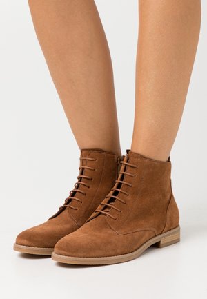 Bottes de cheville en daim marron avec un laçage à l'avant, texture douce, bout arrondi et petit talon empilé. Coutures visibles sur le cuir.