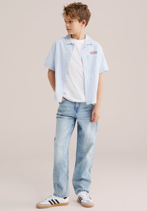 Kiharate poiss, kes kannab helerohelise triibulise lühikese varrukaga särgi valge T-särgi peal, helepesu teksasid ja valgeid tossusid mustade triipudega.