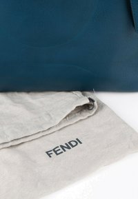 Sac en cuir bleu partiellement visible au-dessus d'un sac anti-poussière en tissu beige plié avec le nom de la marque FENDI imprimé en noir.