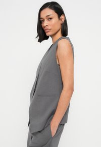 Γκρι αμάνικο σακάκι, εφαρμοστή γραμμή, με βαθύ V-λαιμόκοψη, κουμπιά στο μπροστινό μέρος και πλαϊνές τσέπες. Λείο ύφασμα με διακριτική υφή.