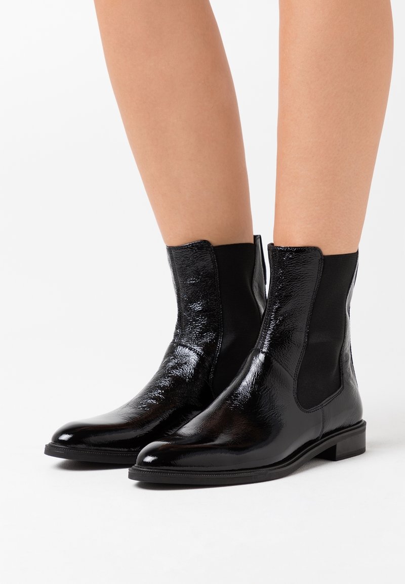 Bottes noires brillantes à la cheville avec panneaux latéraux élastiques portées sur des jambes nues sur un fond blanc.