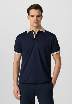 Man draagt een donkerblauw poloshirt met beige kraag en mouwboorden, staand met één hand in de zak tegen een effen lichte achtergrond.