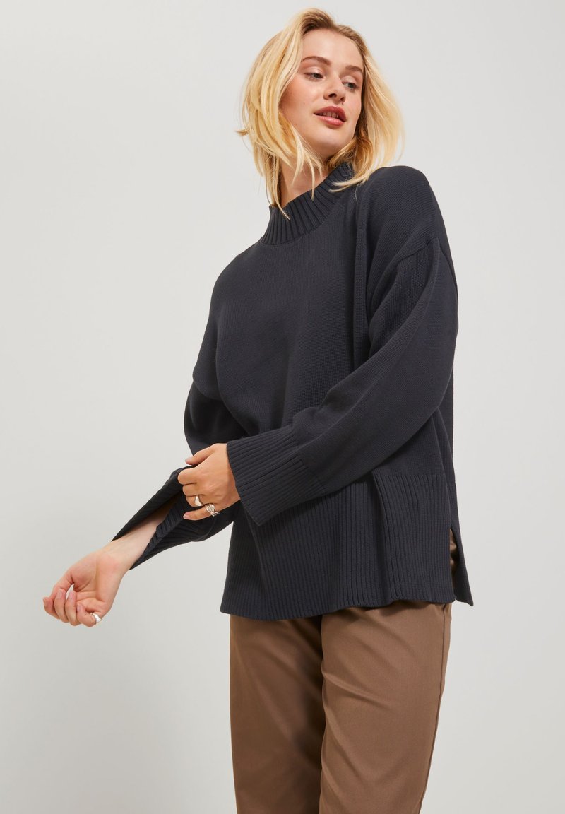 Pull en maille côtelée bleu foncé avec col montant, fentes sur les côtés et manches larges, associé à un pantalon marron clair.
