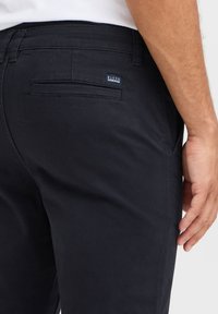 Mörkblå bomullsbyxor med smal passform, utrustade med en bakficka och en liten märkesetikett sydd på midjebandet.