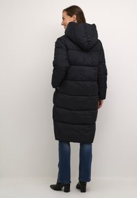 Melna pūkaina jaka ar kapuci, ar quilted dizainu un garu siluetu. Valkāta pār zilām džinsām un melnām papēžu zālēm.