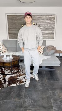 Gray sweatshirt and matching joggers, paired with silver sneakers. Background features a gray couch, patterned pillows, and a circular table.