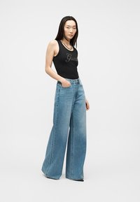 Jeune femme debout, une main dans la poche, portant un haut noir sans manches avec le logo "Guess" et un jean bleu à jambes larges sur fond blanc.
