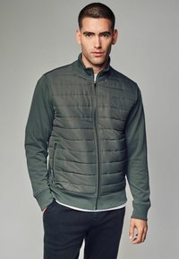 Grüne Zip-Jacke mit gepolstertem Vorderteil, glatten Ärmeln, gerippten Bündchen und Saum; seitliche Taschen; hoher Kragen; über einem hellen Hemd getragen.