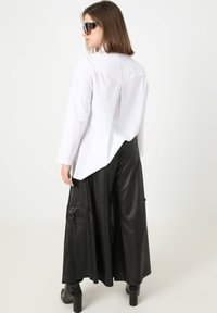 Weißes Langarmhemd mit gespaltenem Rücken, kombiniert mit weiten schwarzen Faux-Lederhosen mit Cargo-Taschen. Schwarze quadratische Sonnenbrille.