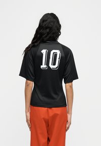 Άτομο με μακριά σκούρα μαλλιά στέκεται γυρισμένο με την πλάτη φορώντας μαύρη αθλητική φανέλα με τον αριθμό "10" σε λευκό στο πίσω μέρος και άνετο κόκκινο παντελόνι.