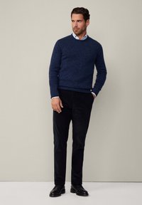 Maglione a maglia blu navy con scollatura a girocollo, abbinato a pantaloni in velluto a coste neri. L'insieme presenta una camicia bianca con colletto sotto.