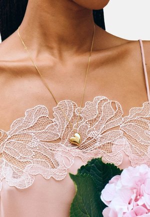 Zelta sirds kaklarota uz smalka rozā mežģīņu topa ar sarežģītu ziedu rakstu. Rozā auduma fons ar zaļām lapām un rozā ziedu.