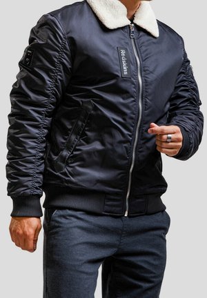 Homme portant une veste bomber noire avec col en polaire blanche et fermeture éclair, associée à un pantalon foncé, sur fond gris uni.
