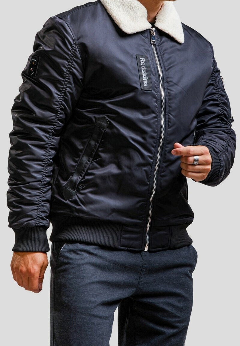 Homme portant une veste bomber noire avec col en polaire blanche et fermeture éclair, associée à un pantalon foncé, sur fond gris uni.