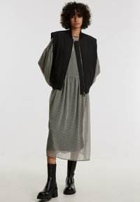 Schwarze ärmellose Bomberjacke über einem beige-karierten, knielangen Kleid, kombiniert mit schwarzen Knöchelboots. Weicher Stoff, lockere Passform und modernes Design.