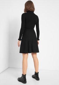 Femme portant une robe noire à manches longues côtelées avec des poignets à volants et des bottes noires montant jusqu'aux chevilles, debout devant un fond blanc.