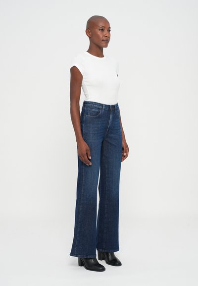 Mujer con cabeza rapada que lleva una blusa blanca de manga corta, jeans de talle alto y pierna ancha de color azul, y botas negras de tacón en el tobillo, de pie sobre un fondo liso.