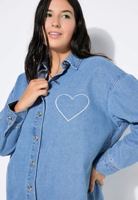 Lichtblauwe denim shirt met een witte hartbroderie op de linkerborst, gouden knopen en een ontspannen pasvorm met lange mouwen.