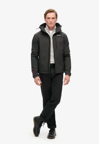 Zwarte geïsoleerde jas met capuchon en geblokt patroon, gedragen over een lichtgrijze trui, gecombineerd met zwarte broek en donkere veterschoenen.