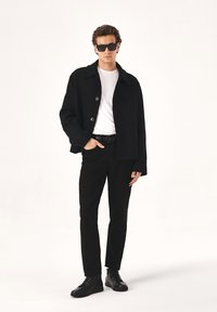 Črn volnen plašč z detajli gumba, bela majica, črne traperice in črne vezene čevlje. Model nosi temna sončna očala. Minimalističen dizajn.