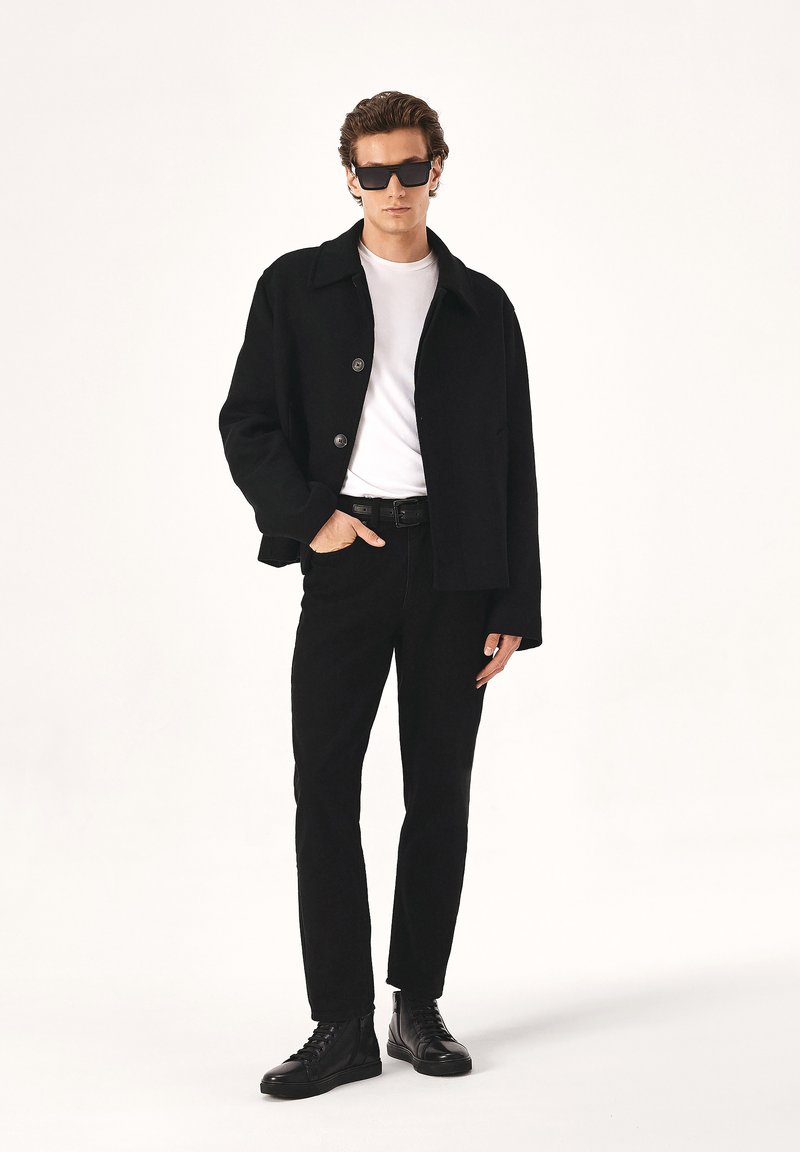 Črn volnen plašč z detajli gumba, bela majica, črne traperice in črne vezene čevlje. Model nosi temna sončna očala. Minimalističen dizajn.