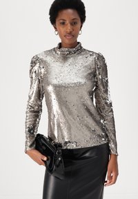 Silberner Pailletten-Oberteil mit langen Ärmeln, hohem Kragen und tailliertem Design, kombiniert mit einer schwarzen, lackierten Clutch und einem Lederrock.