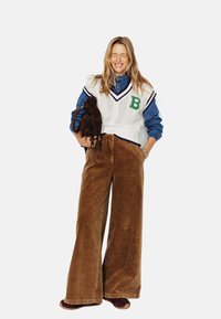 Gilet en tricot blanc avec un "B" vert, chemise en jean bleu, pantalon en velours côtelé marron à jambes larges et chaussures de style mocassin marron. Sac à franges marron.