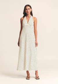 Haljina maxi s halter izrezom od bijelog materijala s crnim točkicama. Glatka tekstura i lepršavi kroj koji naglašava opušteni stil.