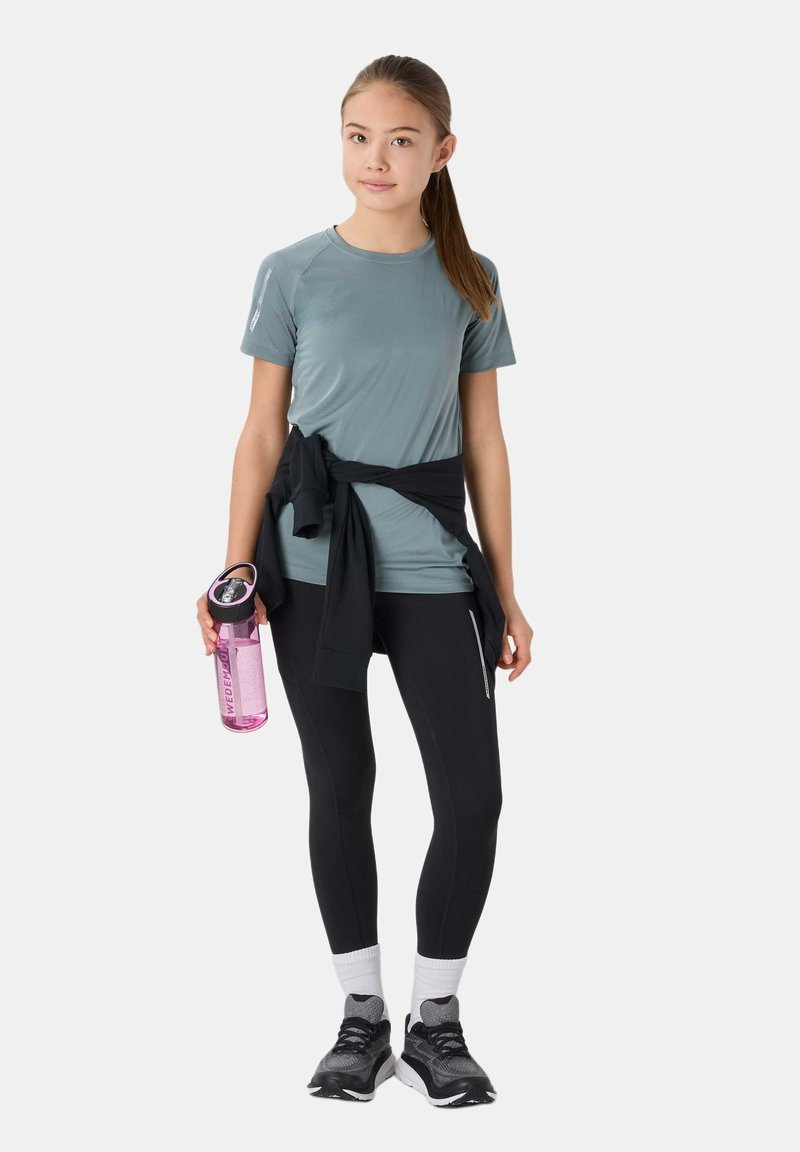 Jeune femme en tenue sportive tenant une bouteille d'eau rose, portant des leggings noirs, un t-shirt gris et une veste noire attachée autour de la taille.