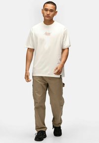 Weißes Baumwoll-T-Shirt mit kurzen Ärmeln, ausgestattet mit einem kleinen orangenen Logo. Kombiniert mit beigen Cargo-Hosen und schwarzen Sneakers.
