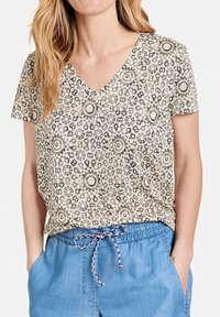 Blusa de manga corta con escote en V en color crema, con intrincado patrón floral en marrón y negro. Tela suave, corte relajado, combinada con pantalones azul con cordón.